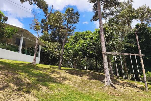 A covered outdoor area with a swing set, surrounded by trees and a white building. - Alsafina Forest Stay (Kemasik)