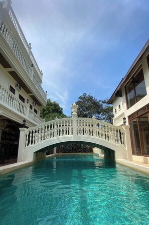 Une piscine surmontée d’un pont à balustrade blanche, un immeuble à plusieurs étages doté de balcons et un ciel d’un bleu limpide.