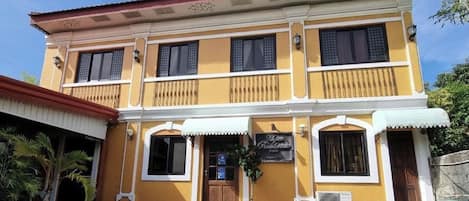 A two-story building with a yellow facade, a sign indicating it is a hotel, and a small garden area with potted plants.