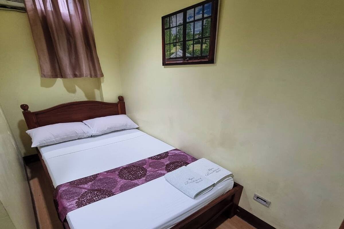 Un lit simple avec une tête de lit en bois, une literie blanche et un coussin décoratif violet. Une fenêtre donnant sur un paysage verdoyant se trouve au-dessus du lit.