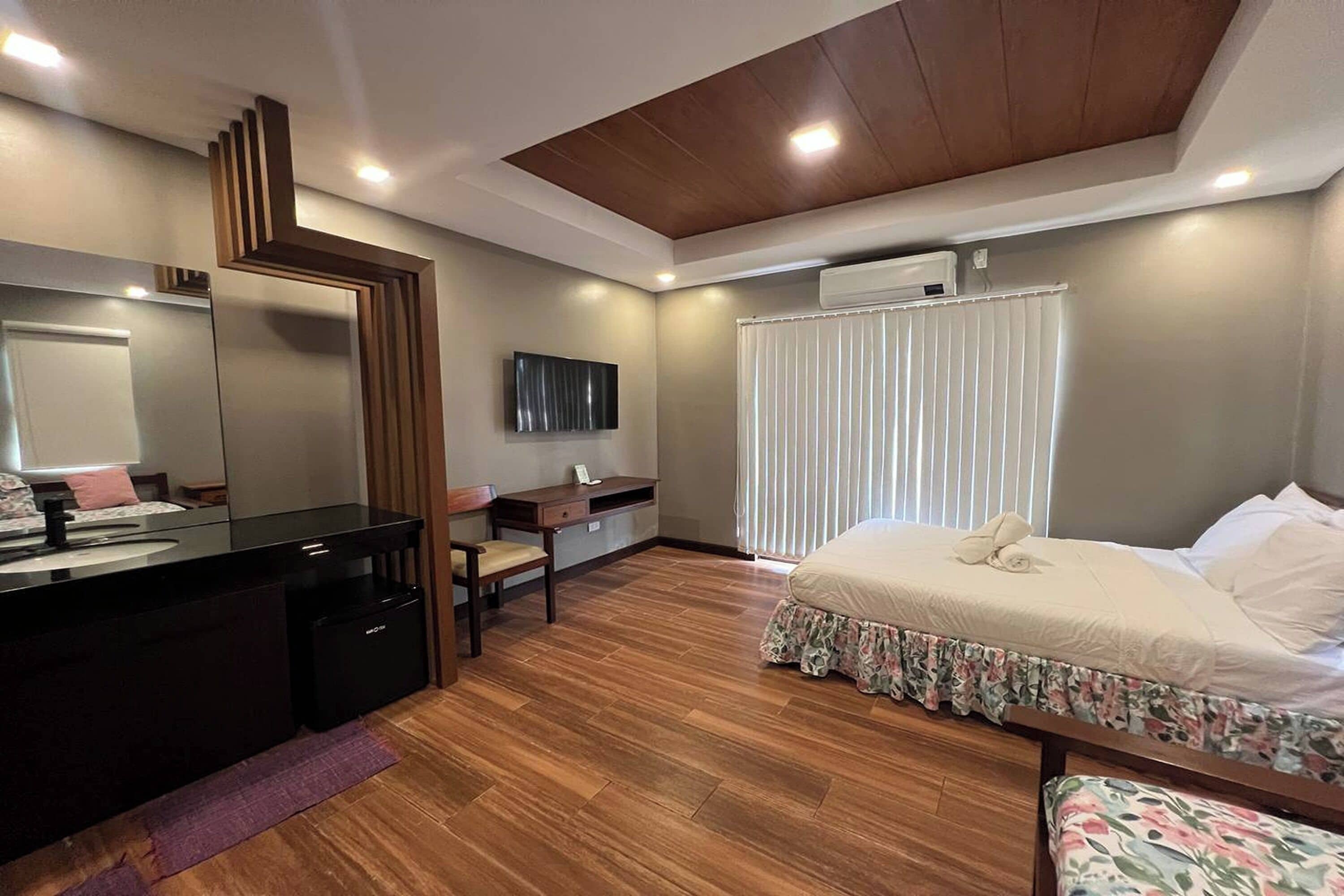 A modern bedroom with a wooden floor, a bed with floral bedding, a desk with a chair, a large mirror, and an air conditioning unit.