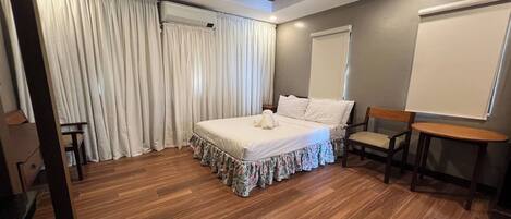 A bedroom with a wooden floor, a bed with a floral bed skirt, a wooden chair, a small table, and a whiteboard on the wall.
