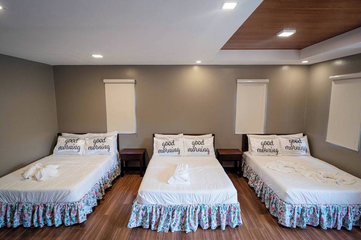 A hotel room with three single beds, each with a floral bed skirt and pillows with 'good morning' text.