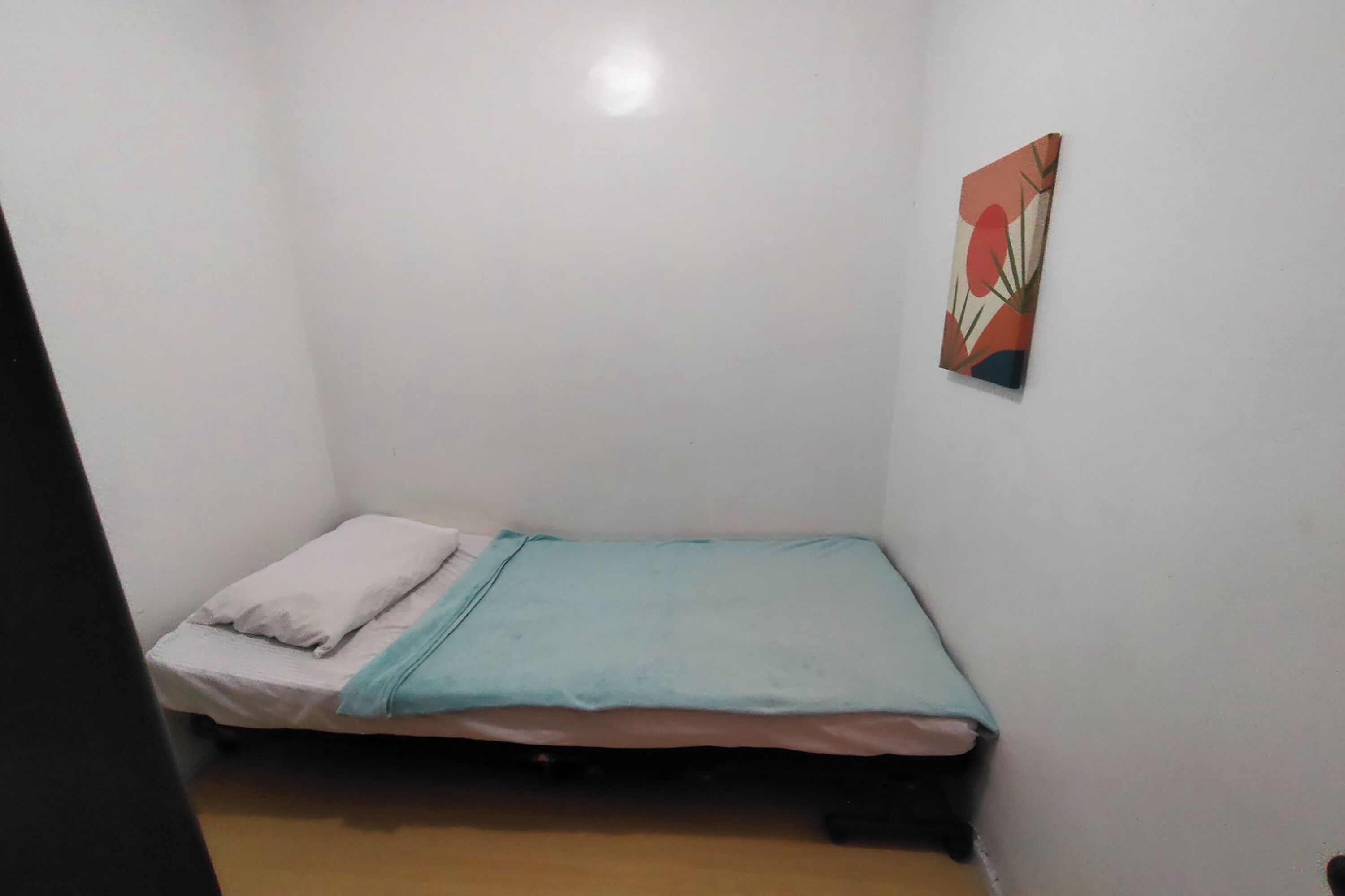 A small, single-bed room with a plain white wall and a colorful abstract painting.