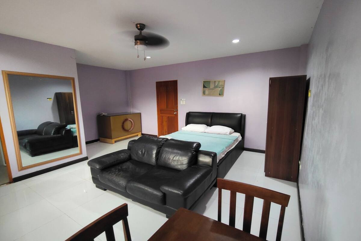 A bedroom with a bed, a black leather sofa, a wooden table, and a ceiling fan.