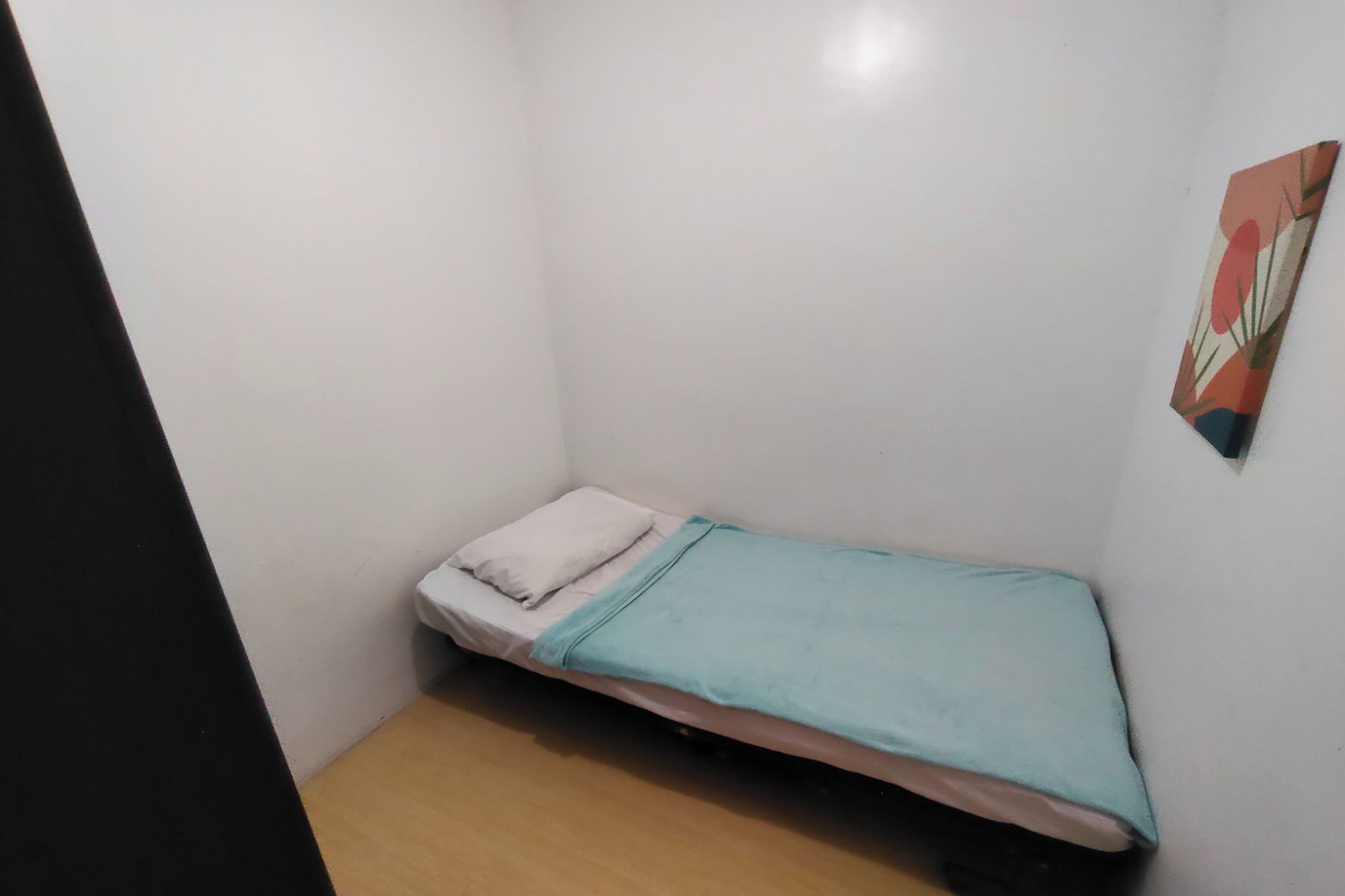 A small, single-bed room with a wooden floor, a white wall, and a colorful floral-patterned artwork on the wall.
