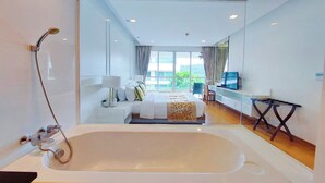 Una habitación de hotel moderna con una bañera grande, una cama con colcha blanca y dorada, un escritorio con silla y un televisor en la pared.