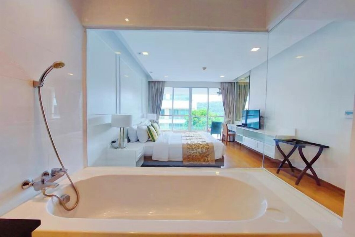 A modern hotel room with a large bathtub, a bed with a white and gold bedspread, a TV on a stand, and a balcony with a view.