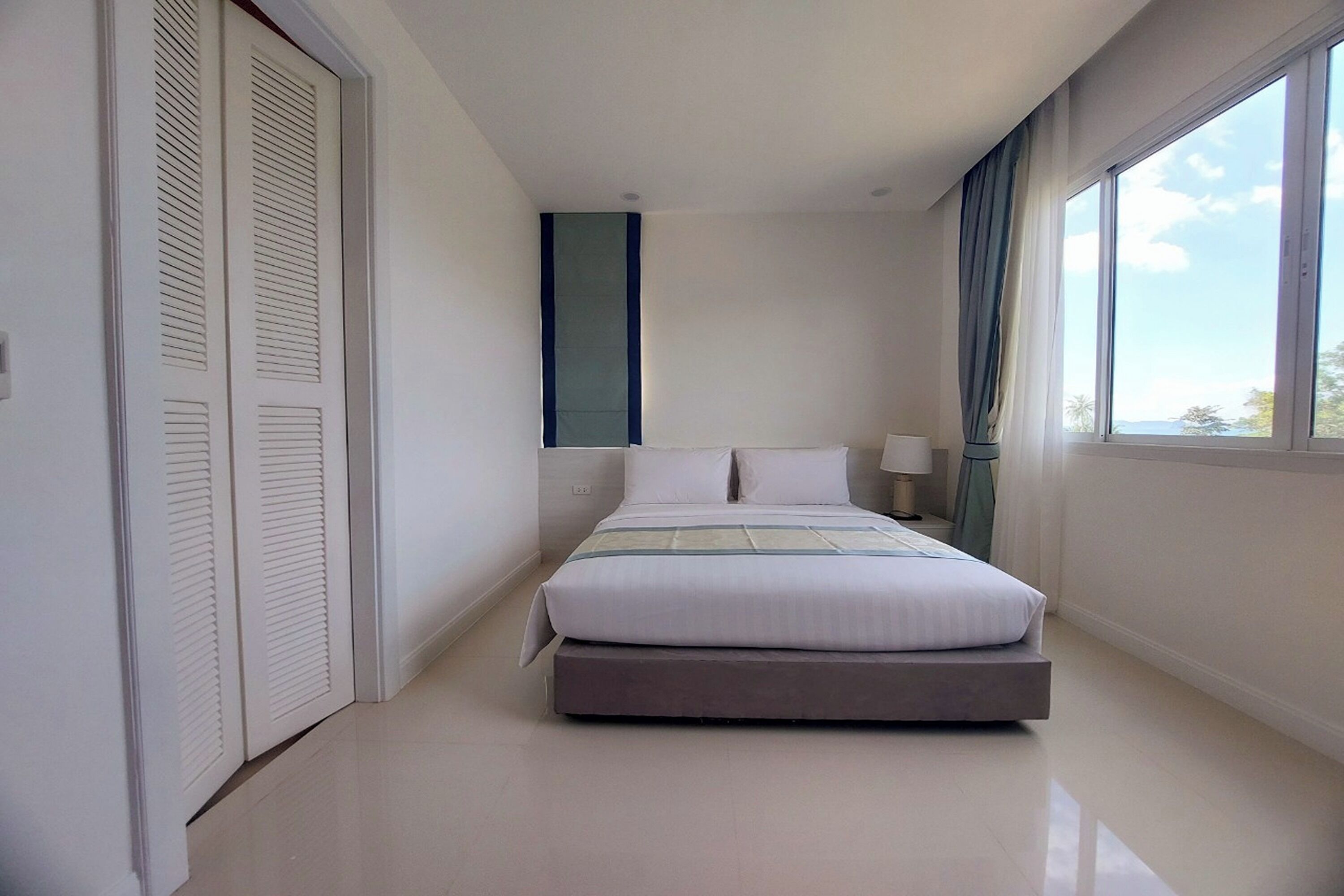 Habitación de hotel moderna con cama, mesita de noche, ventana con cortinas y puerta con paneles persiana.