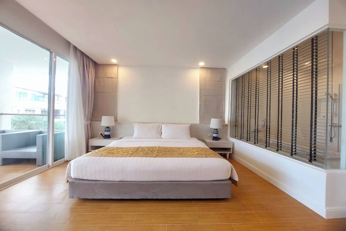 Een moderne hotelkamer met een groot bed, twee nachtkastjes met lampen, een glazen scheiding met jaloezieën en uitzicht op het groen.