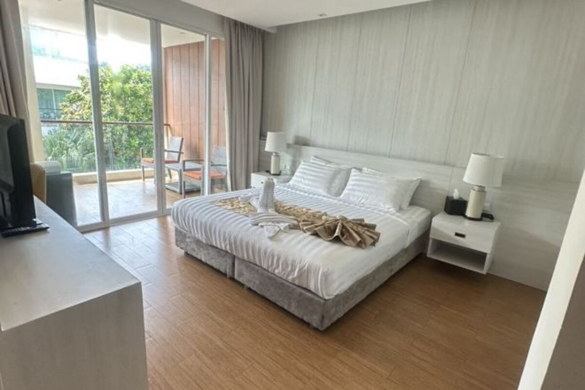 Une chambre d’hôtel moderne avec un grand lit, un téléviseur posé sur un meuble, un balcon avec une chaise et une porte-fenêtre coulissante.