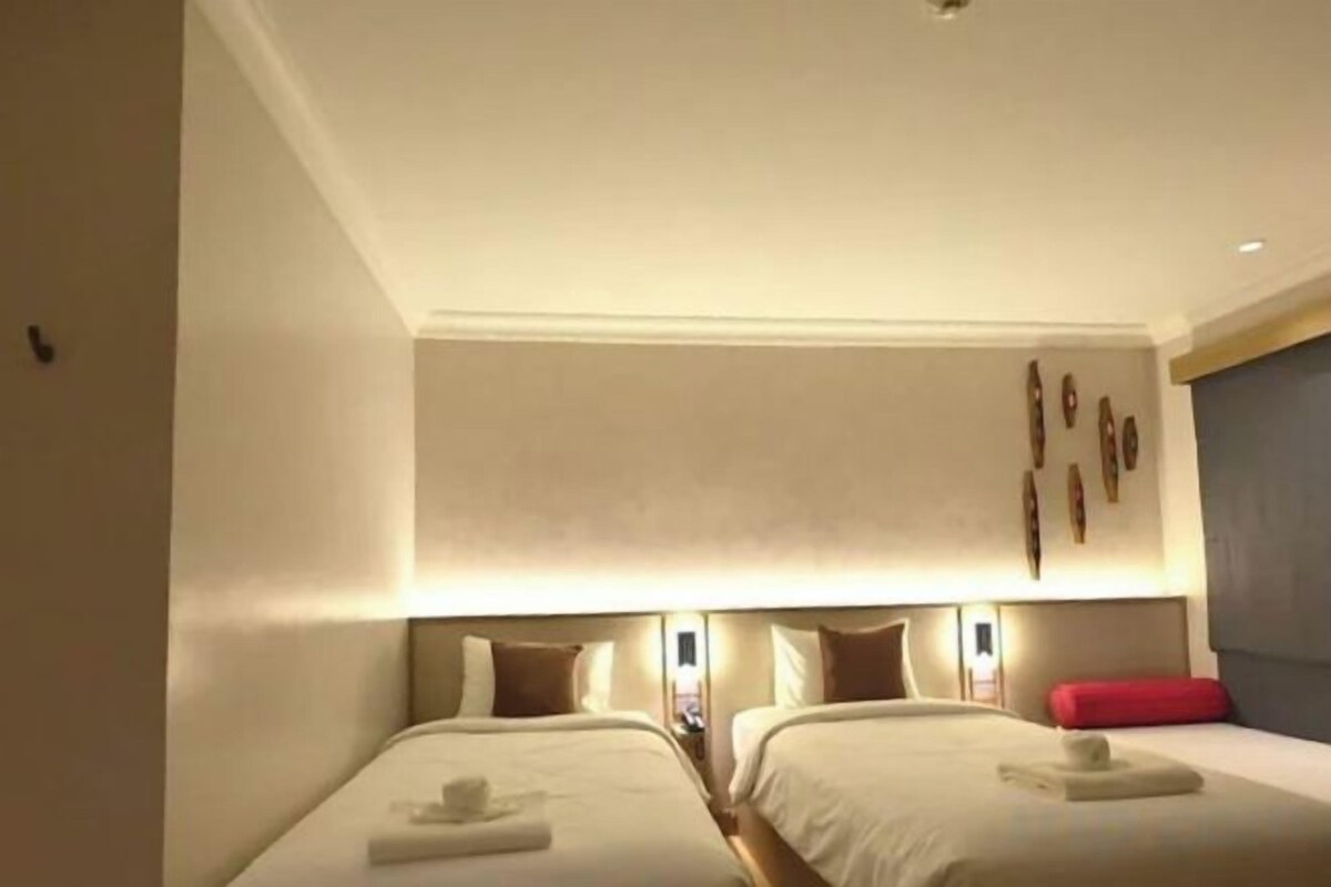 A hotel room with two beds, a red accent pillow, and wall-mounted lighting.