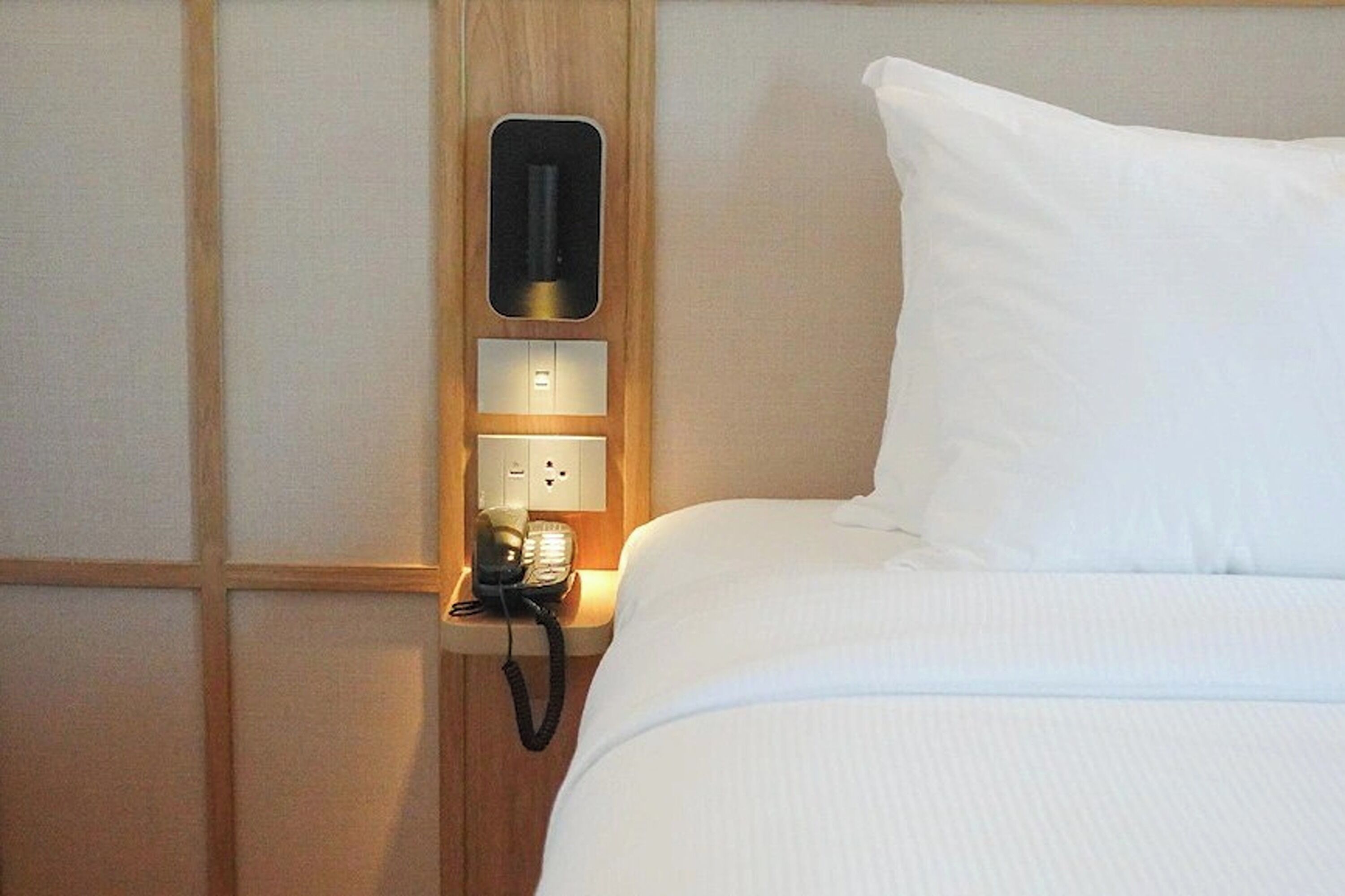 Ein Hotelzimmer mit einem ordentlich bezogenem Bett, einem Nachttischtelefon und einer wandmontierten Bedienkonsole.