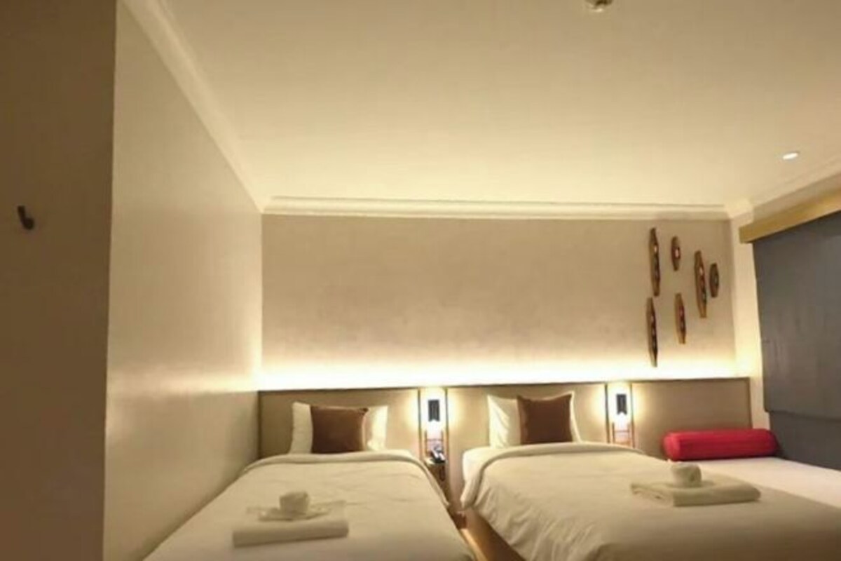 A hotel room with two beds, each with a pillow and a towel. There are wall-mounted lights above the beds and a red pillow on the right side.