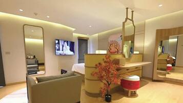 A modern hotel room with a bed, a seating area, a vanity with a mirror, and a television.