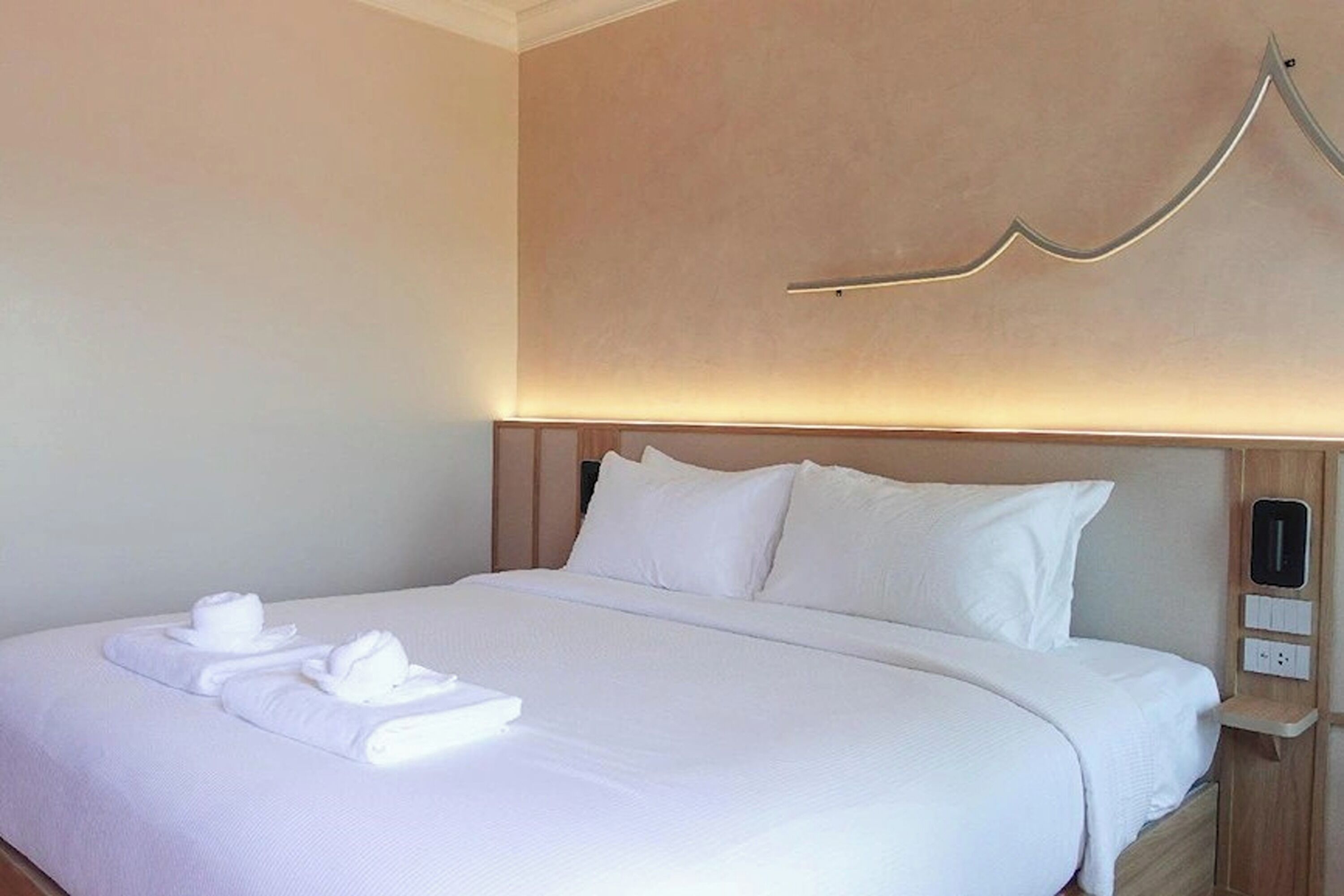 A hotel room with a neatly made bed, white linens, and a modern headboard with a light fixture.