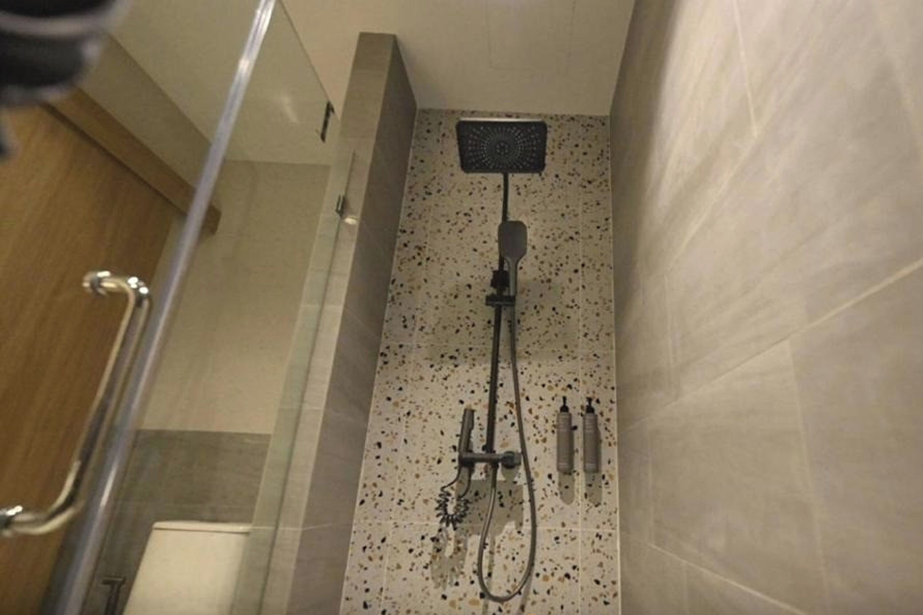 Une douche équipée d’un pommeau de douche à main et d’un robinet mural, avec des murs carrelés et une porte en verre.