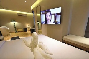 Una habitación de hotel con cama, televisor montado en la pared, mesita de noche y un banco.