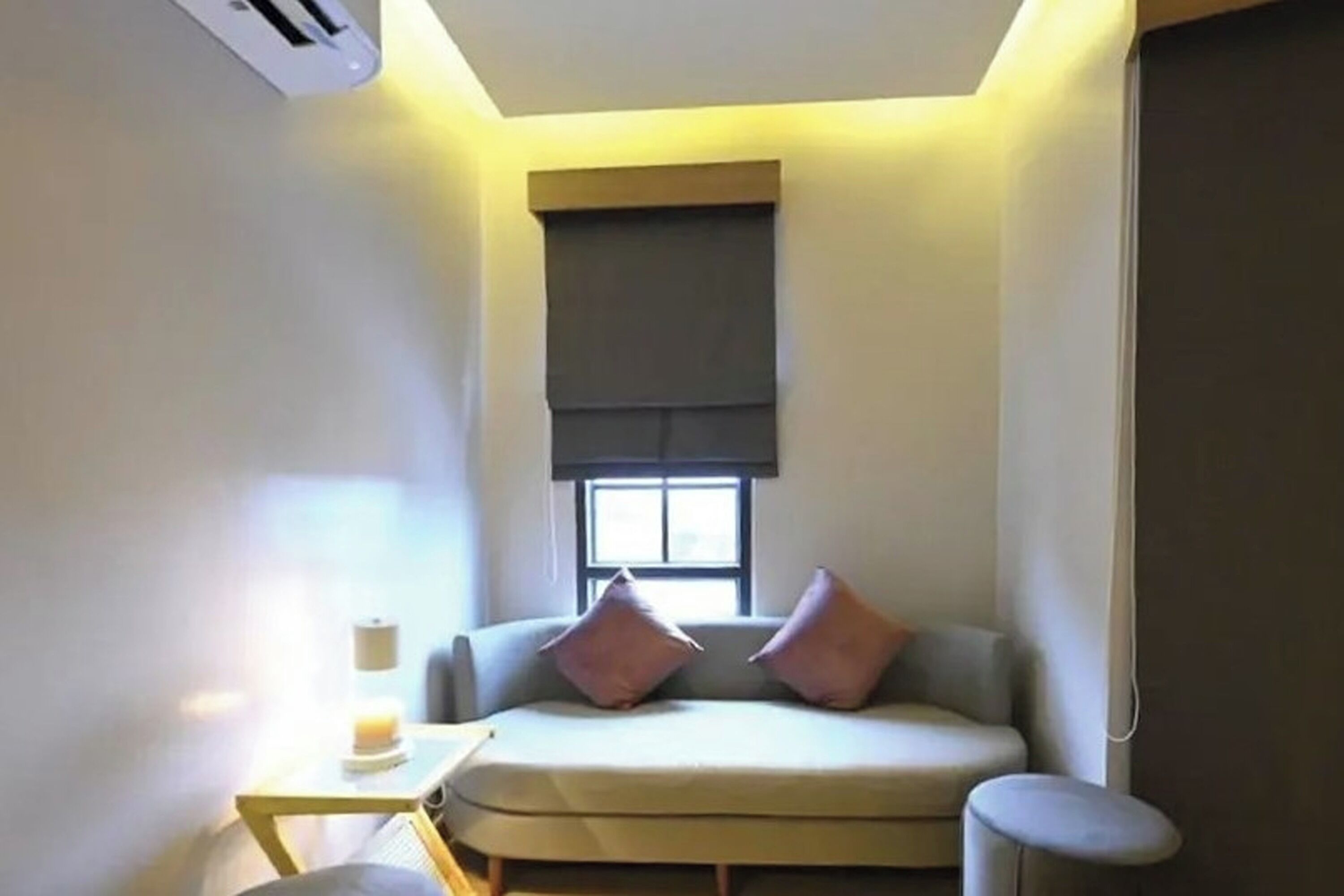A small, cozy room with a sofa, a small table with a lamp, and a window with blinds.