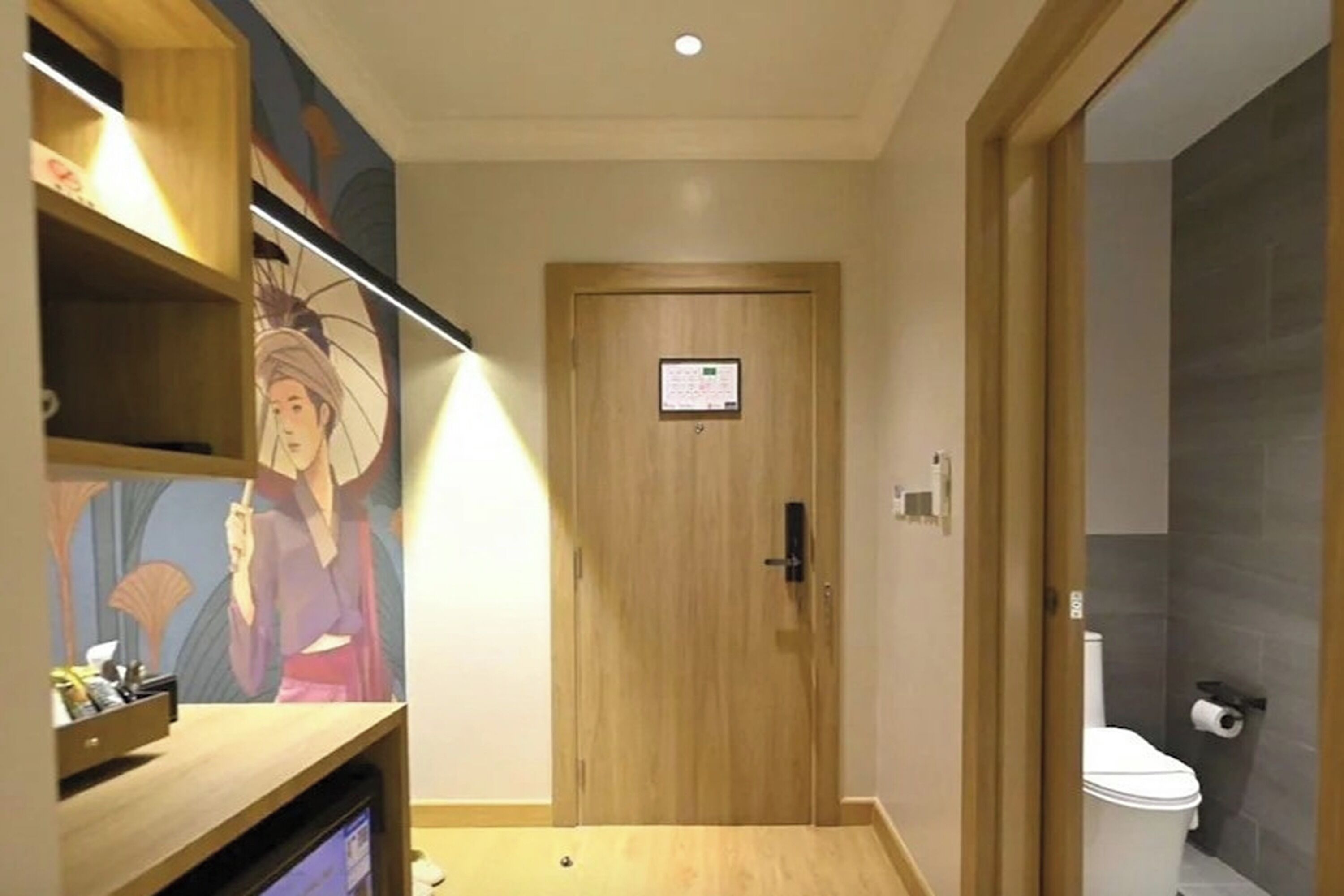 A hotel room with a wooden door, a small shelf with items, and a toilet visible through an open door.