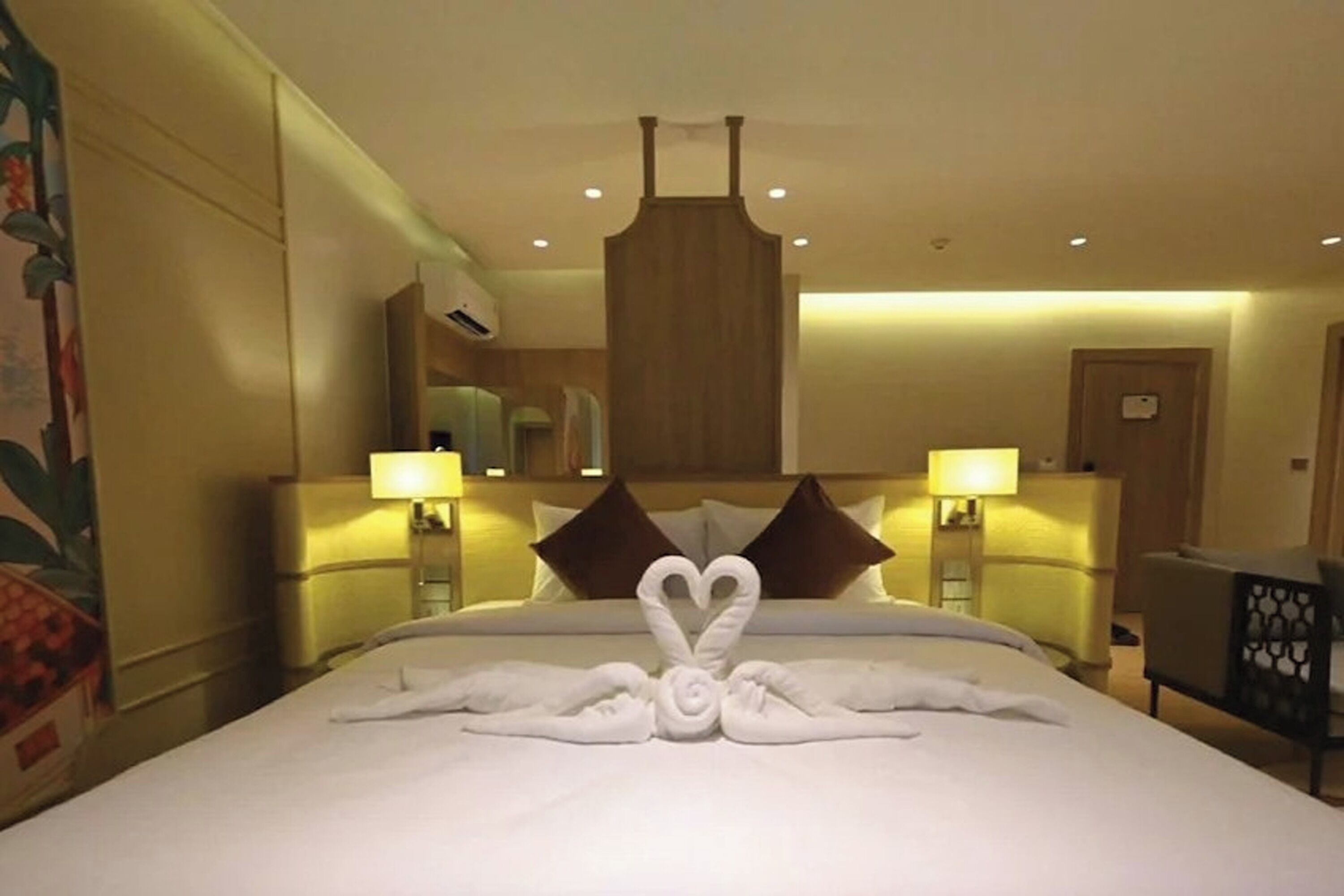 Habitación de hotel con una cama que exhibe una decoración de toallas con forma de corazón y cisne, dos lámparas de noche, un cabecero de madera y una silla con respaldo estampado.