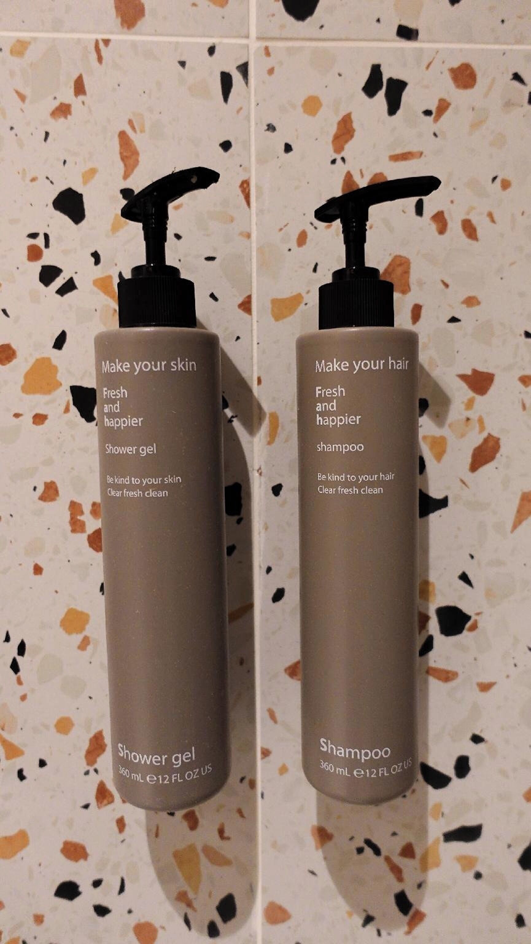 Two bottles of shower gel and shampoo with pumps, labeled 'Make your skin fresh and happier' and 'Make your hair fresh and happier' respectively, against a speckled tile background.