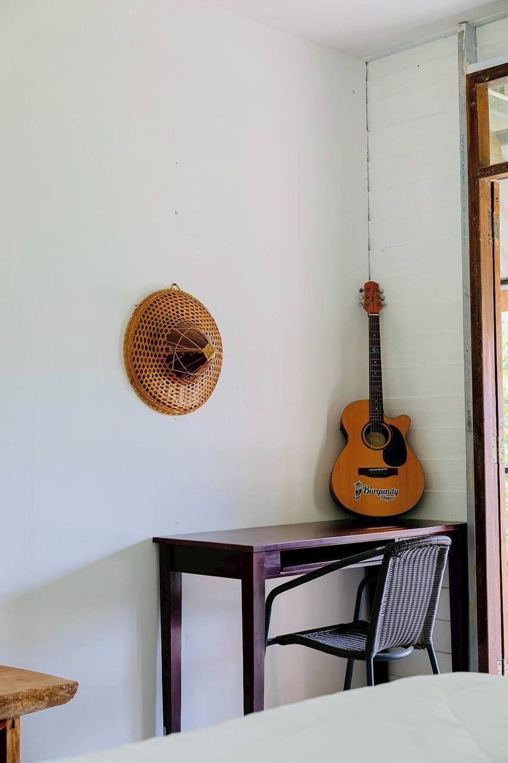 Una habitación con una guitarra apoyada en la pared, una cesta tejida y una pequeña mesa con una silla.