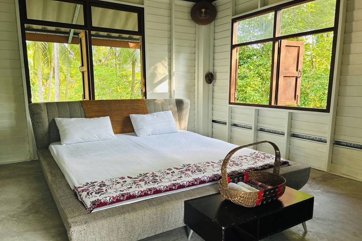 Un lit avec une couvre-lit blanche et une bordure décorative, une tête de lit en bois et une table de chevet noire sur laquelle repose un panier en osier.