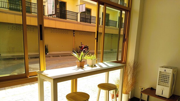 Una habitación luminosa con un ventanal grande, una mesa blanca con dos taburetes y una pequeña estantería con un ventilador.