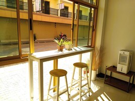 Una habitación luminosa con un ventanal grande, una mesa blanca con dos taburetes y una pequeña estantería con un ventilador.