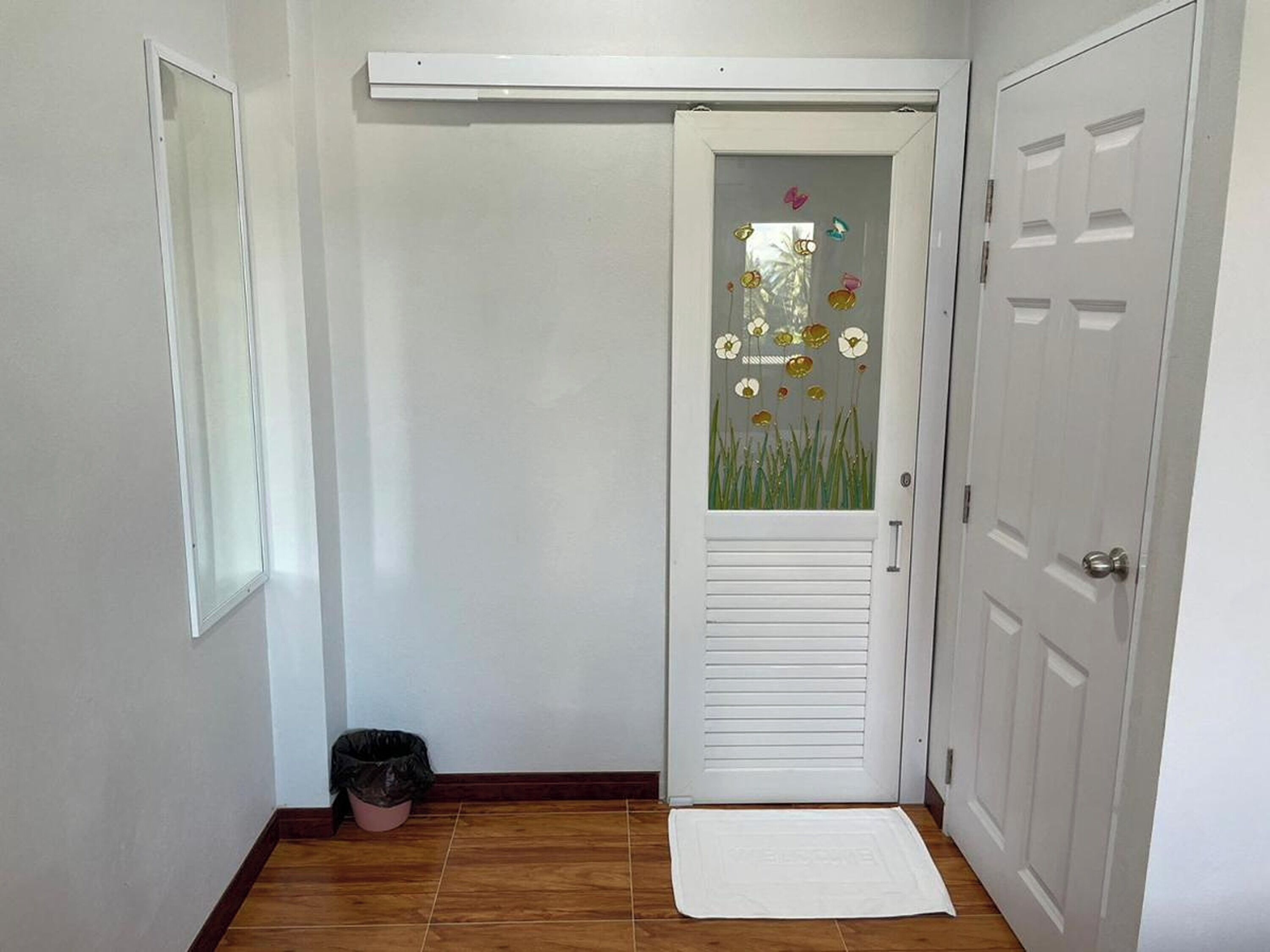 Una habitación con una puerta blanca, un pequeño cubo de basura y un felpudo.