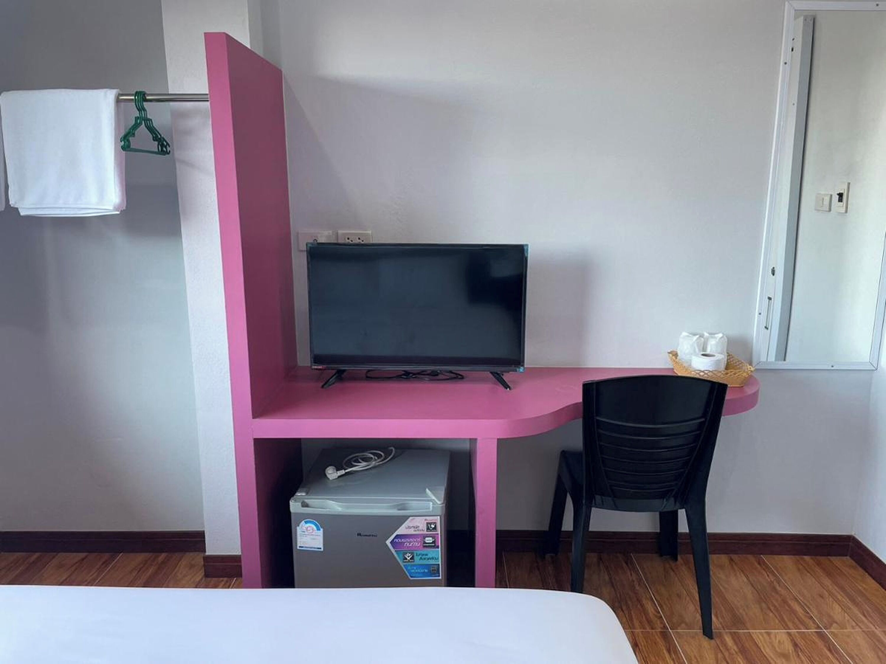 Une pièce avec un bureau rose, une chaise noire, un téléviseur, une serviette blanche accrochée à un cintre et une fenêtre à cadre blanc.