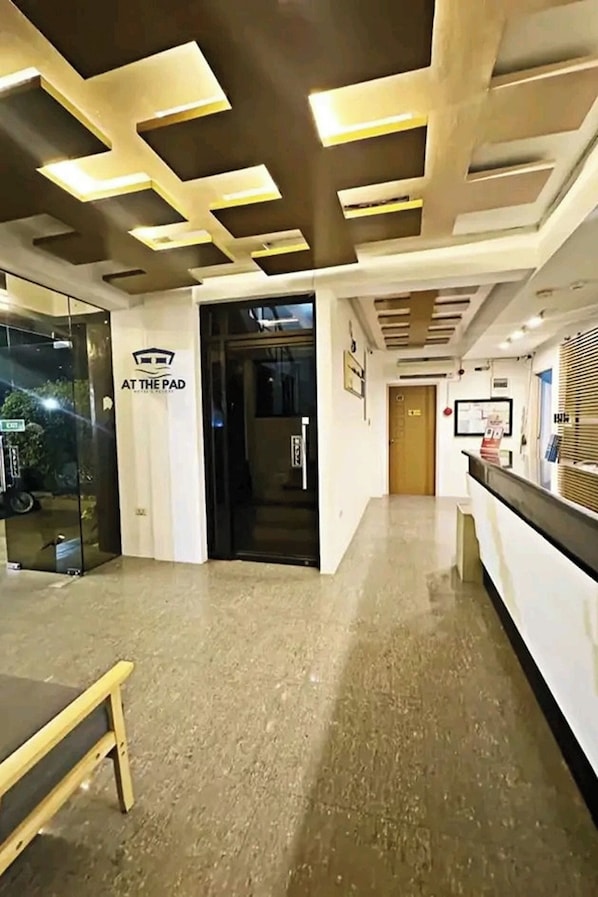 A hotel reception area with a modern design, including a reception desk, a glass door entrance, and a hallway leading to the rooms.