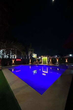 Una piscina illuminata di blu di notte, circondata da un'area verde con palme e un edificio con insegne illuminate.