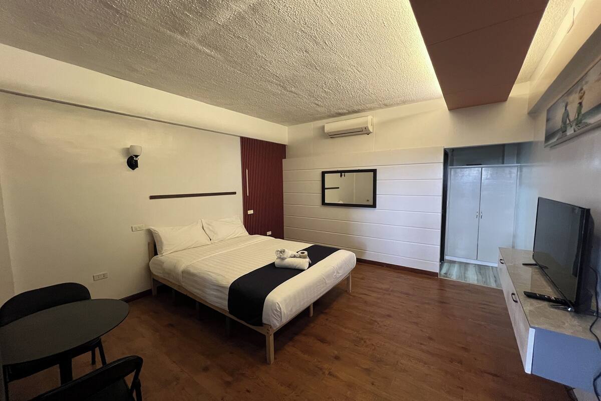Quarto de hotel com cama, escrivaninha, cadeira, televisão e guarda-roupa.
