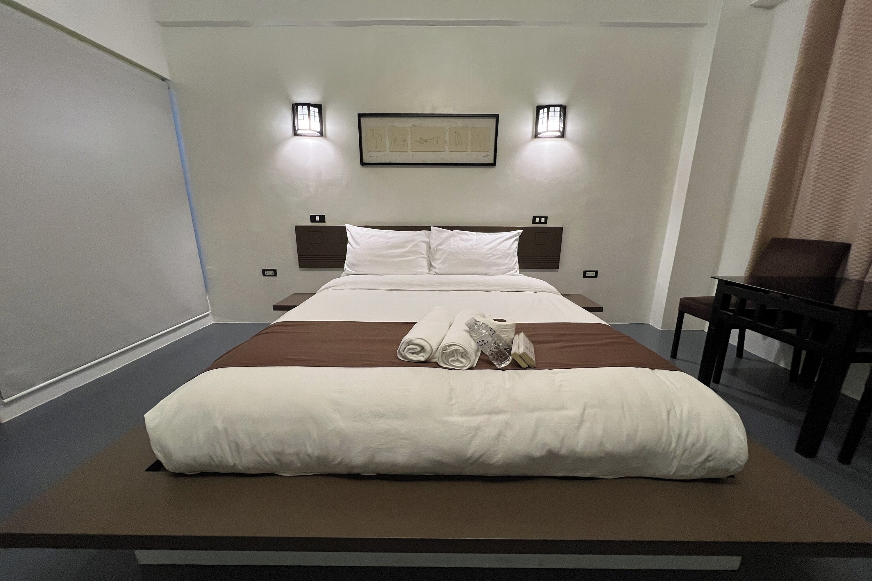 A neatly made bed with white and brown linens, two wall-mounted lights, and a framed artwork above the bed.
