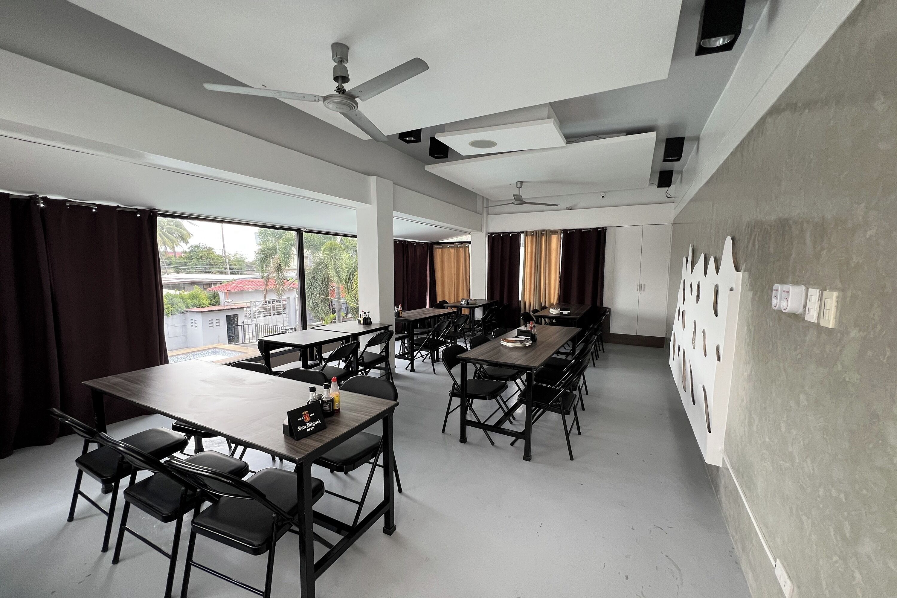 A spacious room with multiple tables and chairs, ceiling fans, and large windows with curtains.