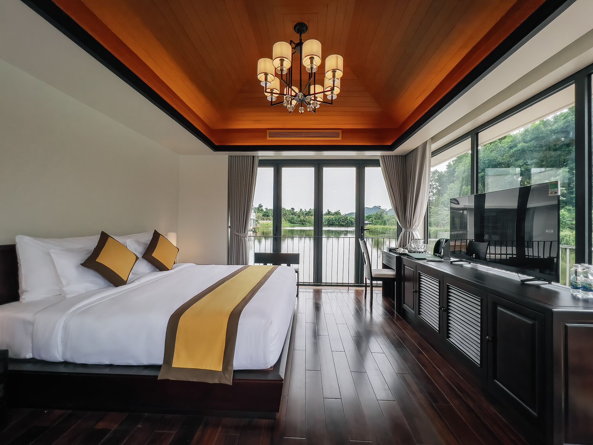A hotel room with a large bed, a desk with a TV, and a balcony with a view of greenery.
