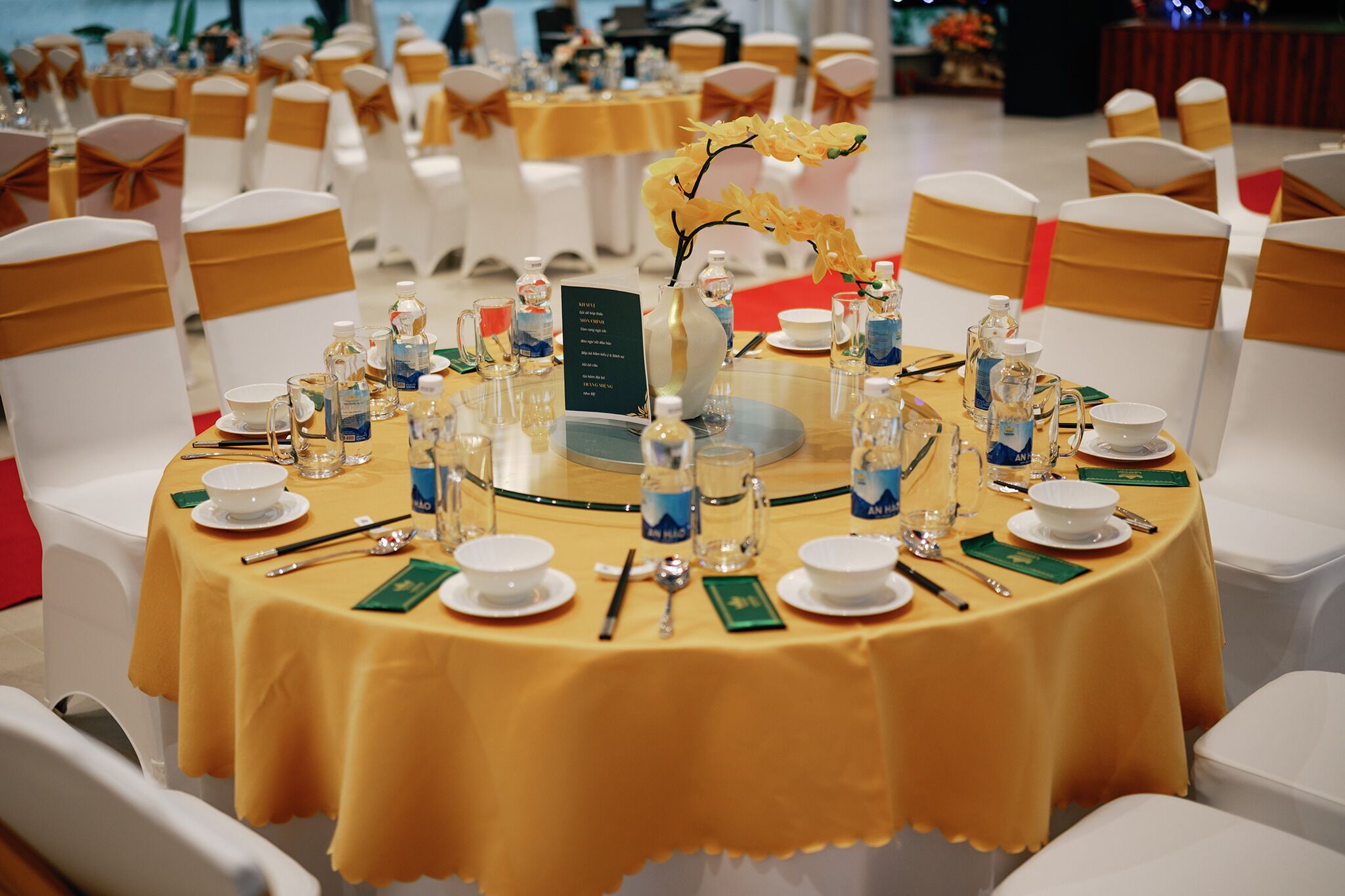 A round table set for a formal event with white chairs and gold sashes, a vase with yellow flowers, and a menu sign.