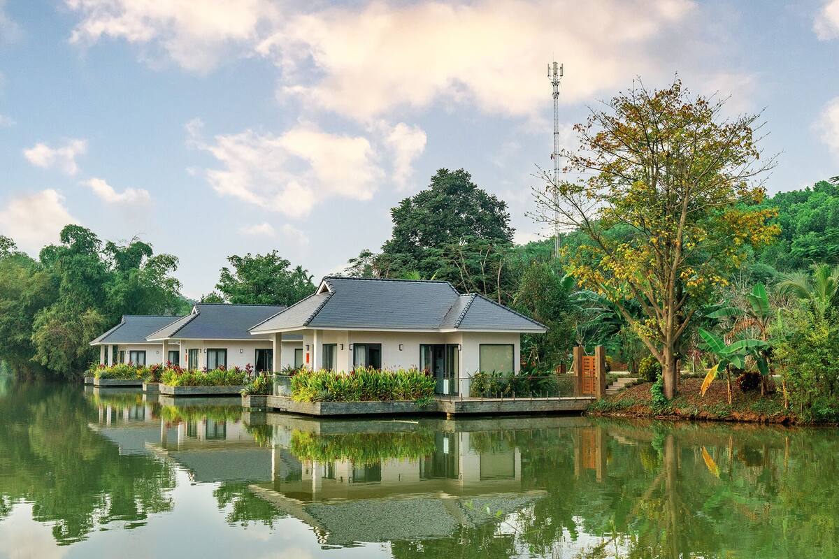 A row of modern, two-story buildings with flat roofs, situated by a calm body of water, surrounded by lush greenery.