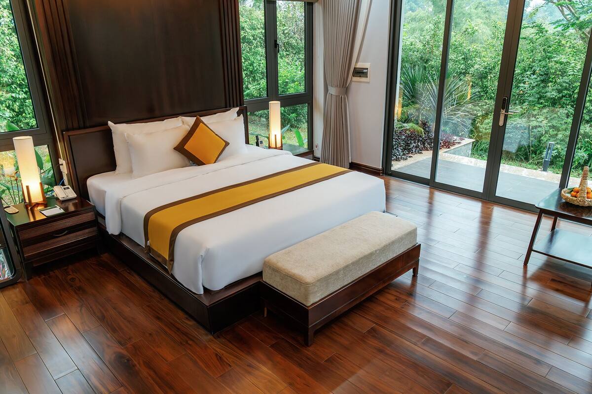A large bed with a wooden headboard, a bedside table with a lamp, and a view of greenery through large windows.