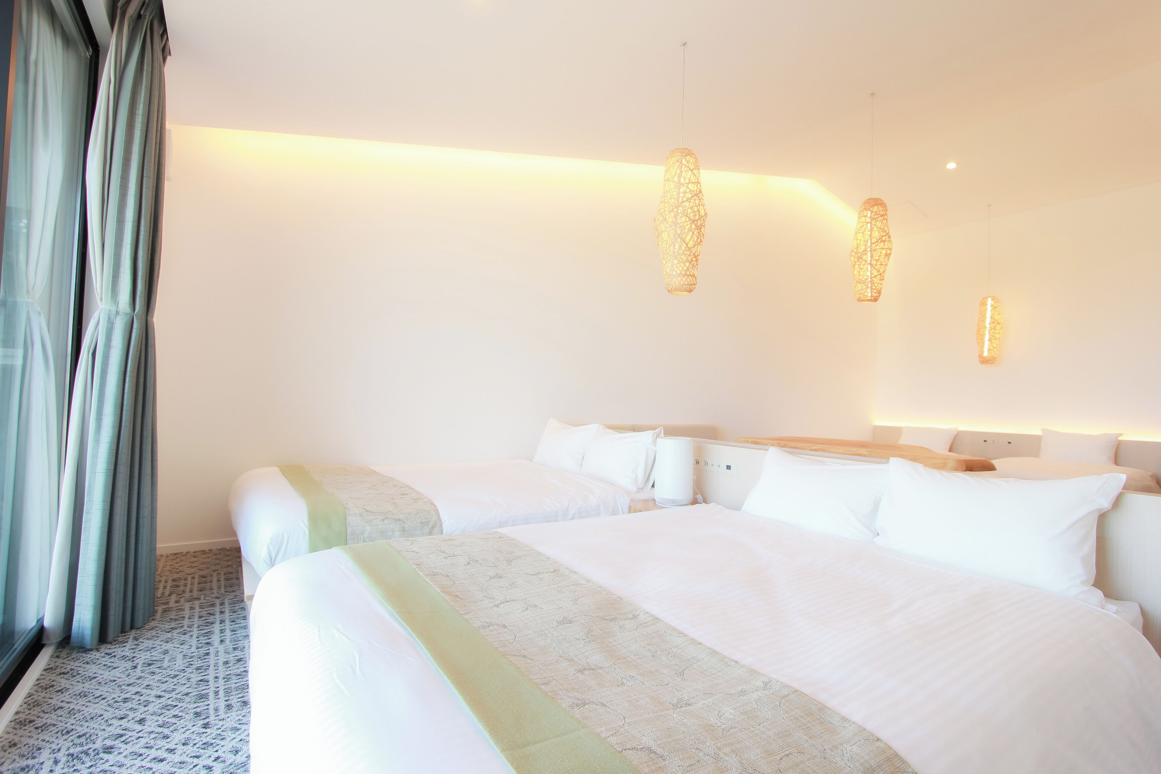 A hotel room with two beds, a large window with curtains, and hanging pendant lights.