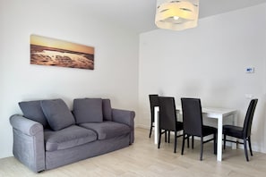 Uma sala de estar moderna com um sofá cinzento, uma mesa de jantar branca com cadeiras pretas e uma luminária pendente.