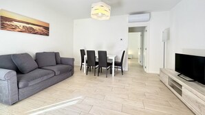 Una sala moderna con un sofá gris, una mesa de comedor con sillas, un televisor sobre un mueble de madera y una lámpara colgante.
