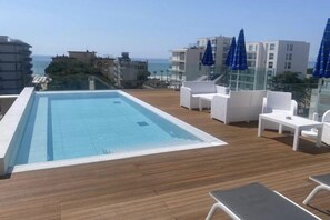 Un espace piscine aménagé sur le toit, offrant une vue dégagée sur l’océan, avec des chaises longues blanches et des parasols bleus.