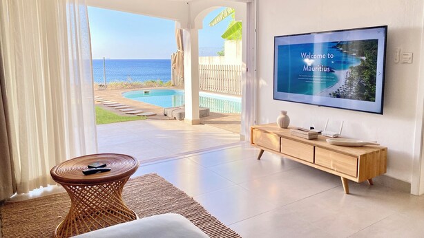 Habitación con televisor mostrando un mensaje de bienvenida, vista a la piscina, una silla de mimbre y un mueble de madera.