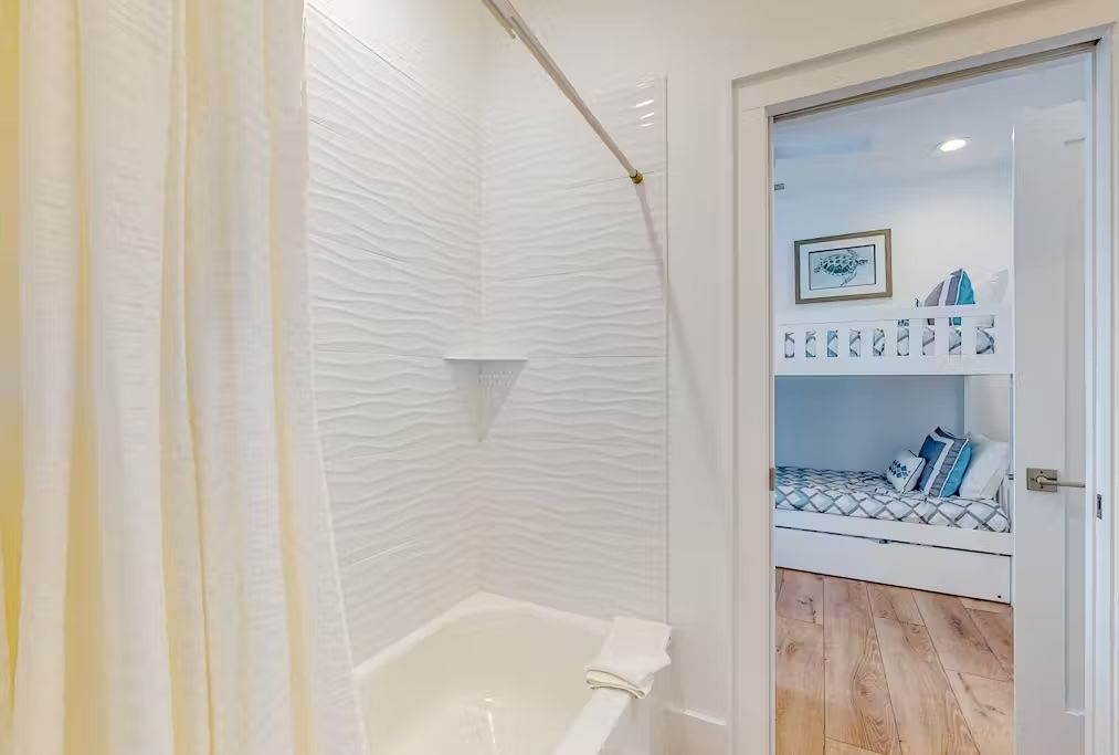 A bathroom with a white bathtub, a shower curtain, and a white textured wall. There is a door leading to a bedroom with a bed and a shelf with decorative items.
