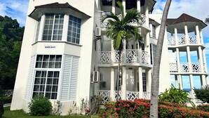 A white building with a balcony, a palm tree, and red flowers.