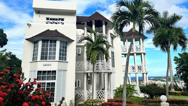 A multi-story building with balconies, a sign reading "BLOCK K0-K7", and surrounding palm trees and red flowers.