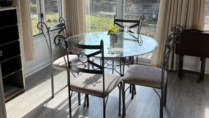 A dining area with a round glass table and metal chairs, large windows with curtains, and a view of a garden.